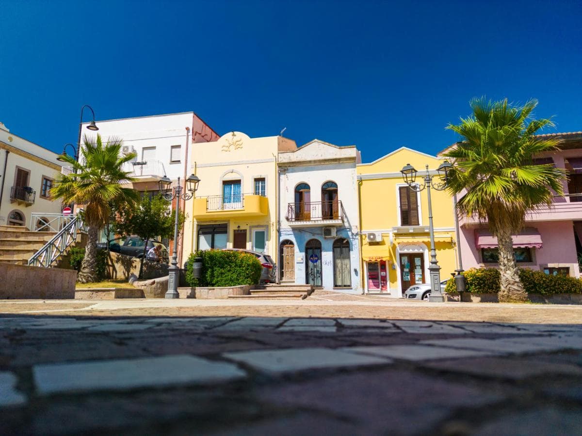 Bunte Häuserzeile mit Palmen an einem Platz in Portoscuso, Sardinien
